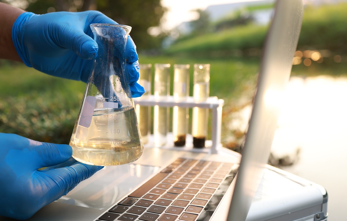 Gloved hands holding a water sample with a laptop, test tubes, and body of water in the background.